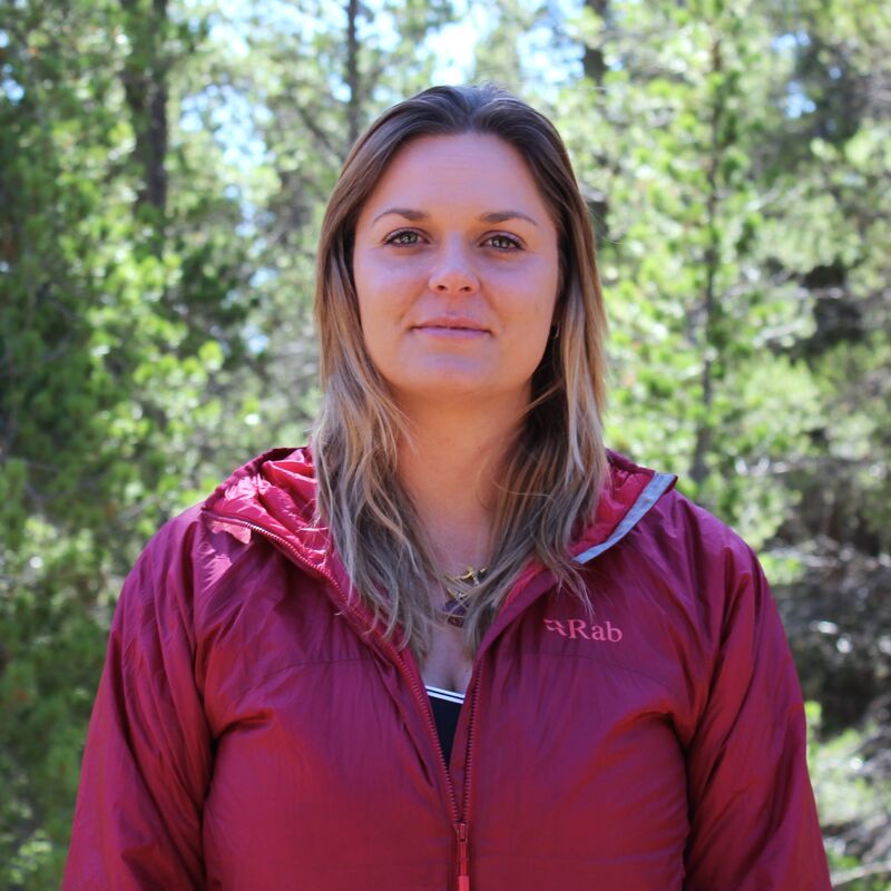 The image shows a woman with long blonde hair, wearing a red hooded jacket. She is standing outdoors, with a background of green trees. The woman is looking directly at the camera with a neutral expression. The lighting is natural, suggesting it is daytime.